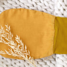 Ein KraftKids Strampelsack Herbst Winter Leinen in senfgelb mit einem dunkleren gelben Bündchen liegt auf einer dicken weißen Strickdecke, akzentuiert durch getrocknetes beiges Gras, das sanft auf das Produkt gelegt wird.