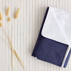 Eine ordentlich gefaltete KraftKids Wickelunterlage Leinen in dunkelblau liegt auf einer weißen Lattenrostfläche neben mehreren getrockneten beigen Grashalmen.