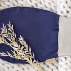 Ein KraftKids Strampelsack Herbst Winter Leinen in Dunkelblau mit beigem Bündchen ruht auf einer dicken weißen Strickdecke, die mit getrockneten Grashalmen bestückt ist. Der Beutel ist mit einem kleinen KRAFTKIDS-Anhänger versehen.