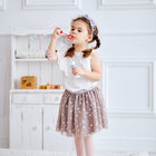 Ein junges Mädchen mit geflochtenen Haaren, weißem Oberteil, gepunktetem Rock und rosa Strumpfhosen pustet Seifenblasen im Haus, während sie auf der KraftKids Krabbeldecke Musselin Zweige braun in einem hellen Raum mit einem kleinen Regal hinter ihr steht.