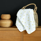 Die KraftKids Babydecke Musselin gelbe Zitronen ist über einen geflochtenen Korb drapiert, der neben zwei gestapelten Weidengefäßen auf einem Holzschrank vor einem schwarzen Hintergrund steht.