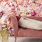 Eine Chaiselongue aus rosafarbenem Samt mit einem KraftKids-Qualitäts-Stillkissen Musselin beige steht neben einem goldenen Beistelltisch mit rosafarbenen und weißen Blumen vor einem farbenfrohen Hintergrund aus rosa, weißen und gelben Rosen.