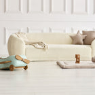 Ein gemütliches cremefarbenes Sofa mit beigen Kissen steht vor einer weiß getäfelten Wand. Auf dem hellen Holzboden liegt die KraftKids Krabbeldecke Musselin beige, weich gefüllt, neben Bauklötzen und einem Plüschhobel in dem hellen, minimalistischen Raum.