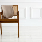 Ein Holzstuhl mit geflochtener Rohrrückenlehne steht auf einem hellen Boden, drapiert mit einer beigen Decke. Die Szene wirkt so gemütlich wie der KraftKids Stillkissenbezug Musselin beige, passend für Theraline 190 cm.