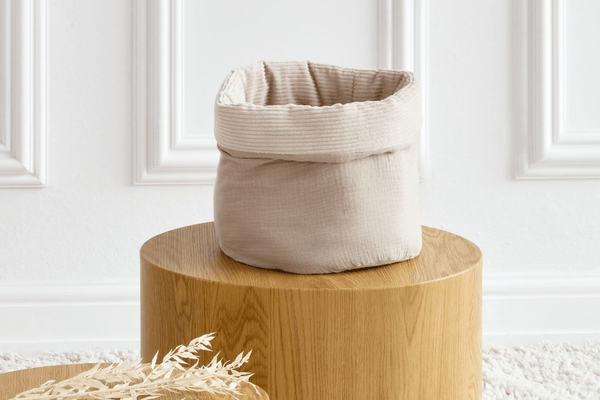 Das KraftKids Körbchen rund Musselin beige hat einen weichen, gefalteten Rand und steht stilvoll auf einem Holzsockel in einem minimalistischen weißen Raum. Getrocknetes Laub in der Nähe macht es ideal als Wickeltisch Organizer oder dekoratives Aufbewahrungskörbchen.