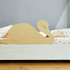 Auf einem weißen Holzbett mit gemustertem Bettzeug steht der KraftKids Rausfallschutz aus Leimholz Schwan, ein kindgerechter Bettschutz in Schwanenform aus hellem Holz, vor einer grauen Wand und einem Holzboden.