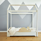 Ein minimalistisches Kinderbett mit einem weißen Rahmen in Hausform ist mit dem KraftKids Rausfallschutz aus Leimholz Schwan als kindgerechtem Schutz und gepunkteter Bettwäsche auf einem Holzboden vor einem grauen, geometrischen Wandbild mit Bergmotiven ausgestattet.