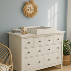 In einem hellen, sonnendurchfluteten Raum sorgen ein KraftKids Wickelaufsatz weiß zum Quer-Wickeln auf einer HEMNES Kommode mit neun Schubladen, ordentlich gefaltete Kleidung, ein Wanddekor mit Löwengesicht, ein Rattanstuhl und eine Topfpflanze für platzsparenden Stil.