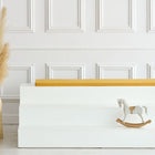 Ein minimalistischer Raum mit einer weiß getäfelten Wand, einer hohen Vase mit Pampasgras, weißen Sitzstufen mit einer KraftKids GmbH Sitzauflage für Ikea TROFAST Leinen in gelb senfgelb und einem kleinen Holzschaukelpferd auf der untersten Stufe.