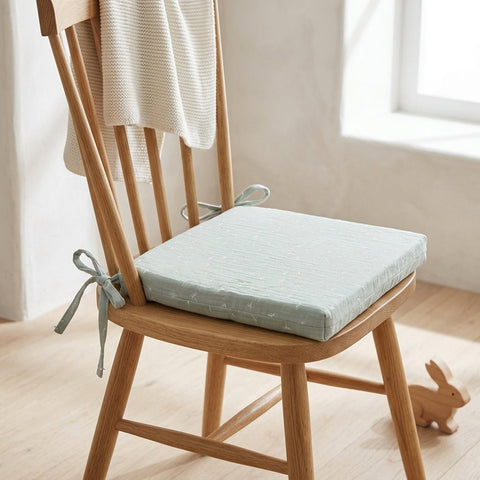 Ein heller Holzstuhl mit einem blassgrünen KraftKids Sitzkissen für Stühle aus Musselin mit abnehmbarem Bezug hat eine beigefarbene Strickdecke über der Rückenlehne und ein kleines Holzspielzeugkaninchen auf dem sonnenbeschienenen Boden daneben.