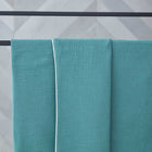 Eine KraftKids Babydecke Musselin nilblau mit weißem Rand ist über einen schwarzen Metallhandtuchhalter drapiert, der vor einer hellgrauen Wand mit Chevron-Muster steht.