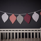 Ein weißes Kinderbett mit einer hellen Matratze steht vor einer dunklen Wand. Darüber hängt die KraftKids Wimpelkette Musselin purpur weiß grau Pusteblumen, die für festliche Farbe sorgt - ideal für Geburtstage.