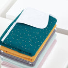 Ein ordentlich gefalteter Stapel KraftKids Wickelunterlage Musselin goldene Punkte auf Petrol liegt auf einer weißen Fläche aus, wobei die oberste Wickelunterlage teilweise aufgeklappt ist und ihre weiße, strukturierte Unterseite zum Vorschein kommt.