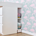 Ein KraftKids Stauraumregal für Wickeltisch in Weiß, passend für 3er oder 4er MALM Kommode, mit offenen Fächern und Drahtkörben voller Kleidung steht vor einer Wand im pinken Flamingo-Design auf Türkis, ergänzt vom Holzfußboden.