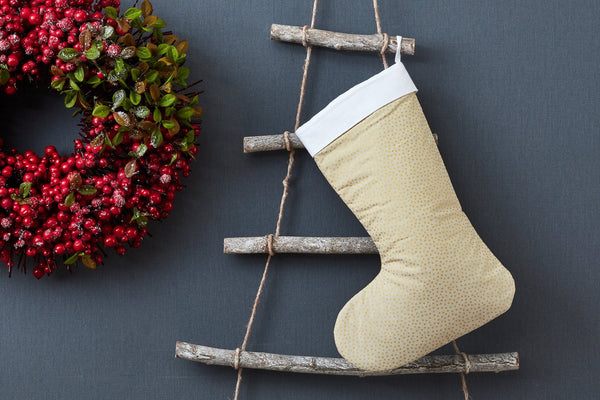 Eine KraftKids Weihnachtssocke mit goldenen unregelmäßigen Punkten auf olivem Grün hängt an einer rustikalen Holzleiter vor grauer Wand. Links ergänzt ein grüner Kranz mit roten Beeren die festliche Kinderzimmerdeko.