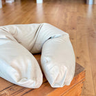 Ein hellbraunes U-förmiges qualitäts Stillkissen Leinen Natur Braun von KraftKids liegt auf einer Holzfläche, mit einem Holzboden und weißen Säulen im Hintergrund.