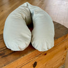 Ein leicht grünes weißes, ergonomisch designtes Qualitäts Stillkissen Leinen von KraftKids liegt auf einer Holztruhe, mit Holzboden im Hintergrund.