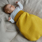 Ein Baby liegt auf einer beigefarbenen Decke, trägt ein weißes Oberteil und einen senfgelben KraftKids Strampelsack Frühling Sommer Waffel Piqué Schlafsack. Weiche, sternförmige Kissen liegen daneben.