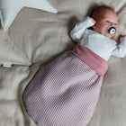 Ein Baby mit Schnuller liegt auf einer beigen Decke, eingekuschelt in den KraftKids Strampelsack Herbst Winter Waffel Piqué rosa. In Weiß gekleidet und von sternförmigen Kissen umgeben, wirkt die Szene gemütlich und verträumt.