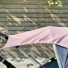 Ein KraftKids Sonnensegel Musselin goldene Punkte auf Rosa ist als Schattenspender über ein Kinderwagenverdeck drapiert, das im Freien durch einen Holzzaun mit Sonnenlicht und Blätterschatten dargestellt wird.