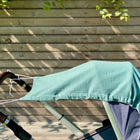 Ein Kinderwagen mit braunen Griffen wird von dem KraftKids Sonnensegel Musselin nilblau beschattet. Er steht im Freien im hellen Sonnenlicht, in der Nähe eines Holzzauns mit grünen Blättern über ihm.