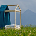 Ein minimalistisches Bettgestell aus Holz mit einer blaugrünen Decke und einem KraftKids Bettbaldachin Musselin goldene Punkte auf Petrol, das über eine Seite drapiert ist, steht auf einer grünen Wiese mit Wildblumen vor nebelverhangenen Bergen unter einem wolkenverhangenen Himmel.