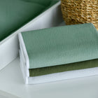 Ein Stapel von drei ordentlich gefalteten Handtüchern in Grün, Dunkeloliv und Weiß liegt neben einer KraftKids Wickelunterlage Doppelkrepp Grün Jade, mit einem geflochtenen Korb und einem grünen Stoffartikel im Hintergrund.