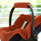 Eine Nahaufnahme einer schwarzen Babyschale mit einer KraftKids Babydecke für Babyschale Sommer Doppelkrepp Rot Herbstrot und einer Griffhülle, die in einem Innenraum in der Nähe eines Fensters mit horizontalen Jalousien aufgestellt ist.