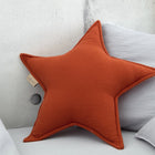 Ein KraftKids Sternkissen Doppelkrepp Rot Herbstrot in Sternform setzt einen gemütlichen, modernen Akzent, wenn es vor einem hellgrauen Kissen und einer Betonwand platziert wird - ideal, um jedem Kinderzimmer Charme zu verleihen.