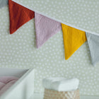 Eine Nahaufnahme der KraftKids Wimpelkette Doppelkrepp Grau Gelb Ros Herbstrot, die an einer weißen gepunkteten Wand über einem Kinderbett mit einer rosa Decke und einem gewebten Behälter hängt - perfekt für die Kinderzimmer-Deko.