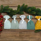 Fünf kleine KraftKids Adventskalender Musselin Gelb Grün Grau Tüten in Grün, Grau und Gelb mit weißen Bändern werden auf Holz mit Tannenzweigen und roten Beeren präsentiert und bilden eine festliche Weihnachtsszene.