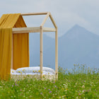 Ein minimalistisches Hausbett mit dem KraftKids Bettbaldachin Musselin goldene Punkte auf Gelb und weißer Bettwäsche steht in einer Wildblumenwiese vor Bergen unter bewölktem Himmel.