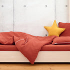 Ein ordentlich gemachtes Bett mit KraftKids Bettwäscheset Doppelkrepp Rot Herbstrot, einem passenden Kissen und einem gelben Sternkissen vor einer hellen Betonwand und einem Holzboden - ideal für einen erholsamen Schlaf in 100% Baumwolle.