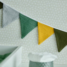Eine KraftKids Wimpelkette Doppelkrepp Grün Gelb Grau Herbstgrün hängt an einer hellen Wand mit weißen Tupfen - eine ideale Kinderzimmer-Deko. Im Vordergrund liegen eine grüne Decke und ein Behälter mit weißem Deckel.